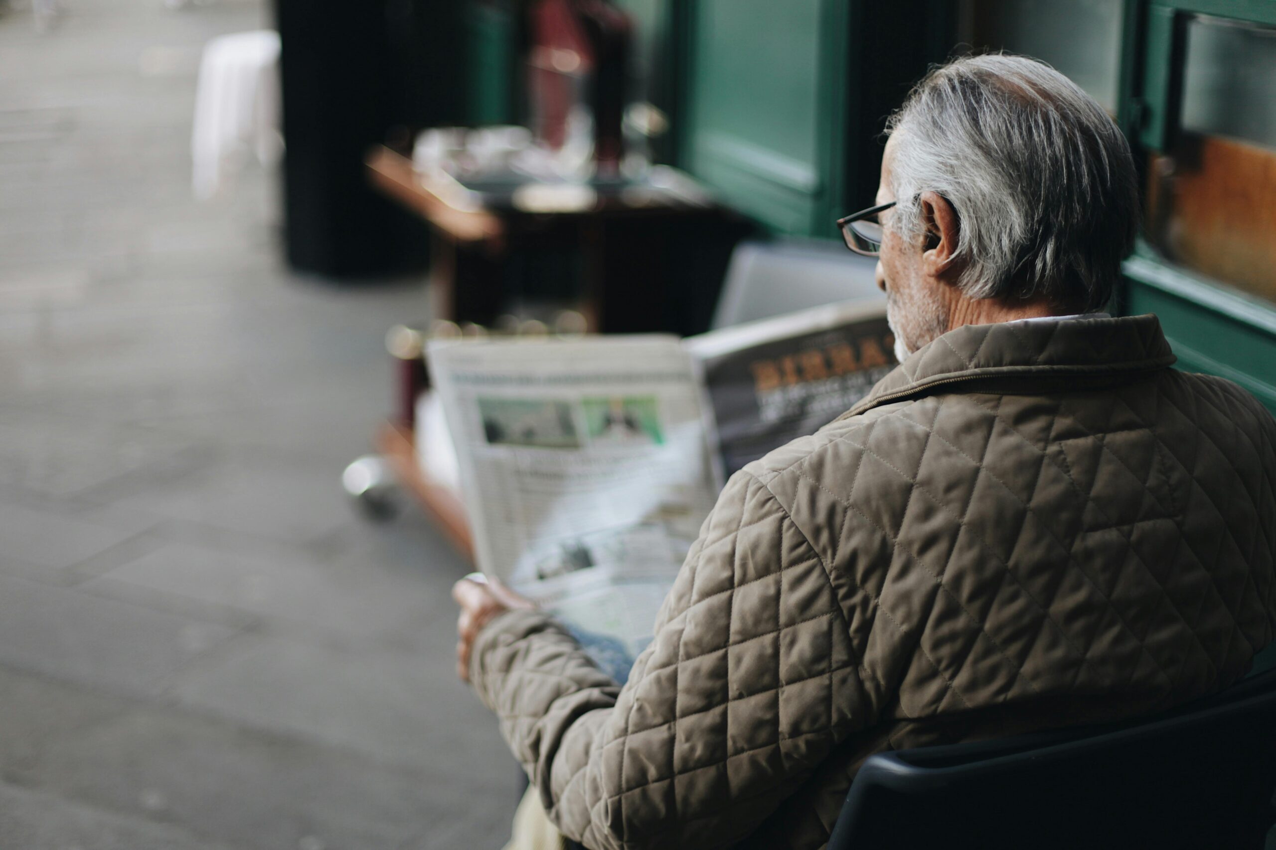 Elderly man reading a newspaper in an outdoor cafe setting, enjoying a peaceful moment.