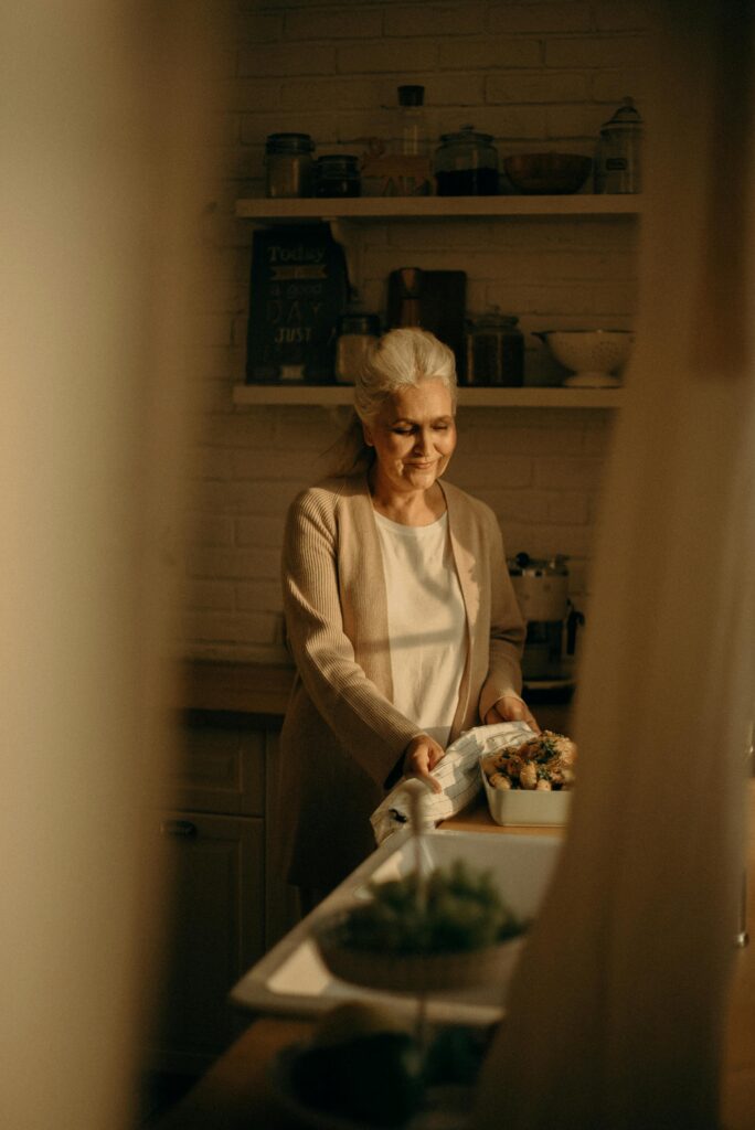 Senior woman with gray hair preparing a meal in a warmly lit kitchen setting.