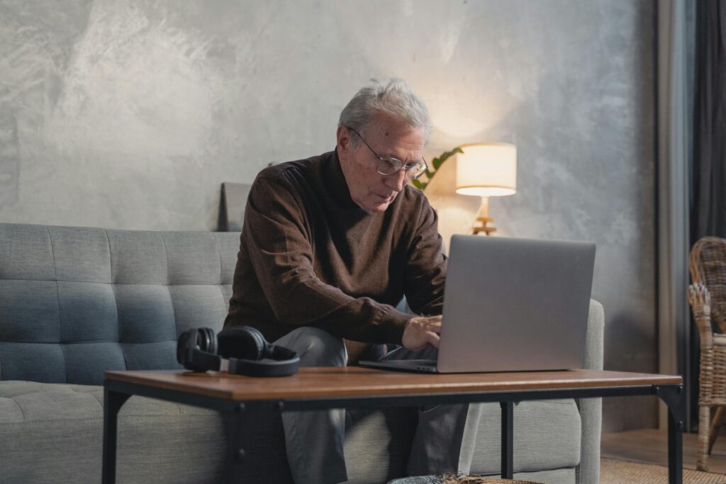 Elderly man using a laptop at home, working on online tasks comfortably from his sofa.
